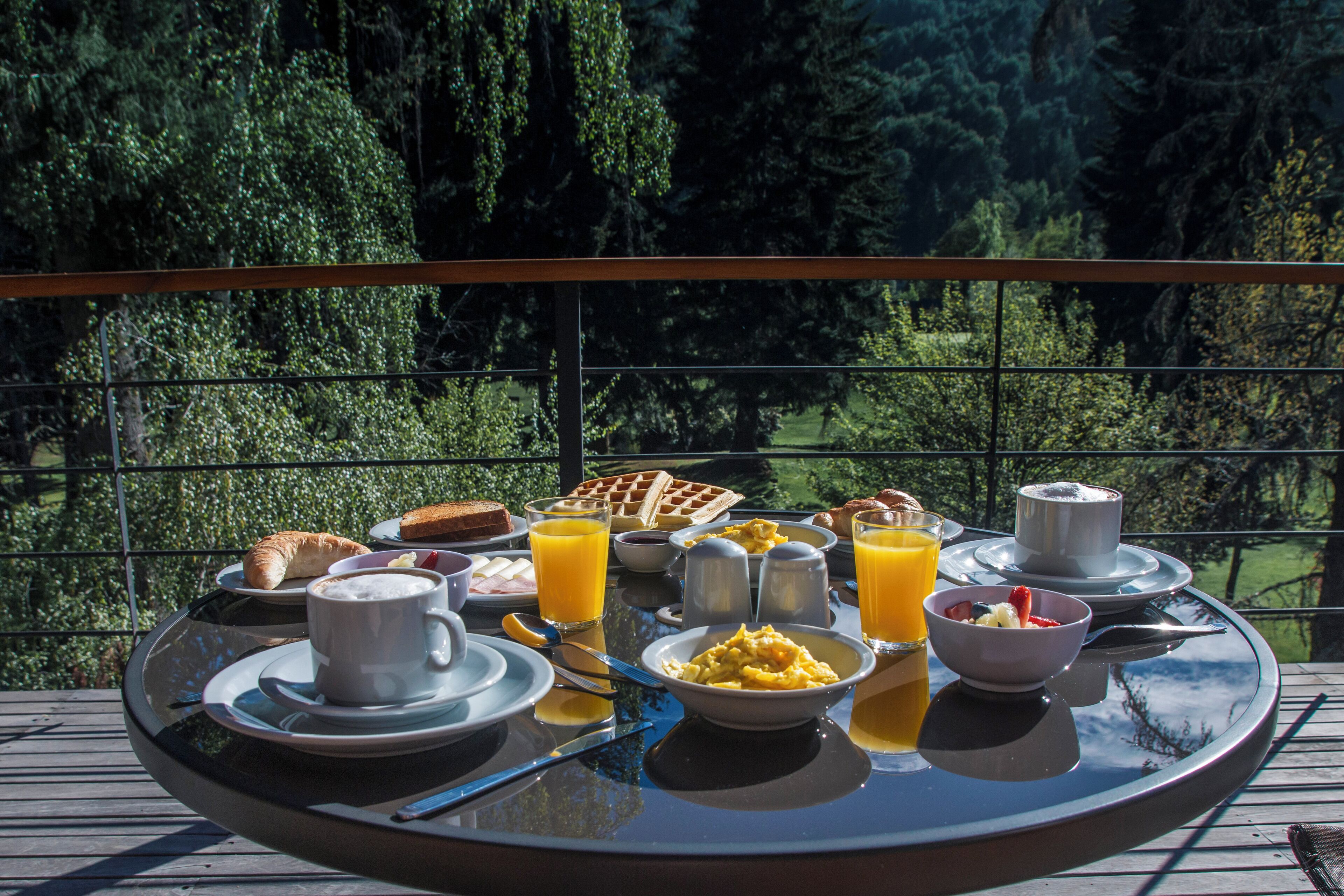 Ein Frühstückstisch mit Croissants, Waffeln, Rühreiern und Orangensaft auf einem Balkon mit Blick auf einen Wald.