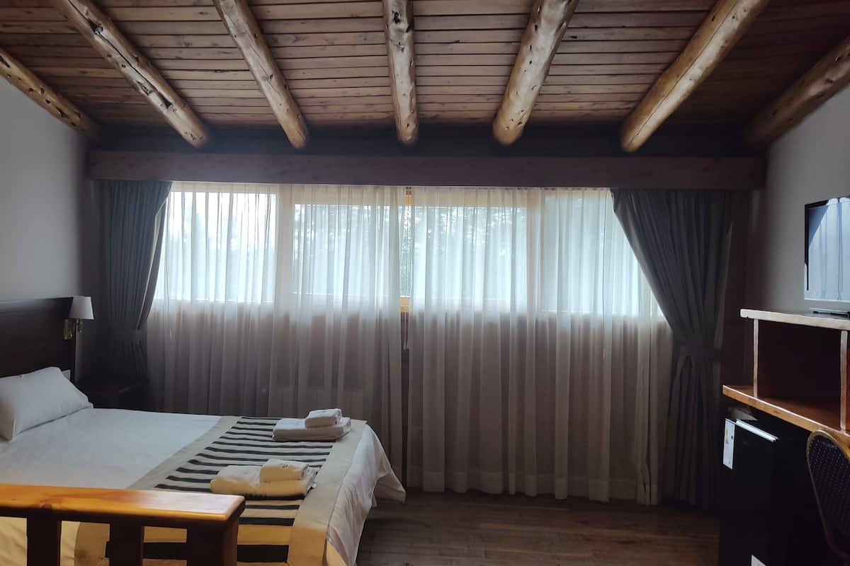 A hotel room with a wooden ceiling, a bed with white linens, a TV on a wooden stand, and a window with sheer curtains.