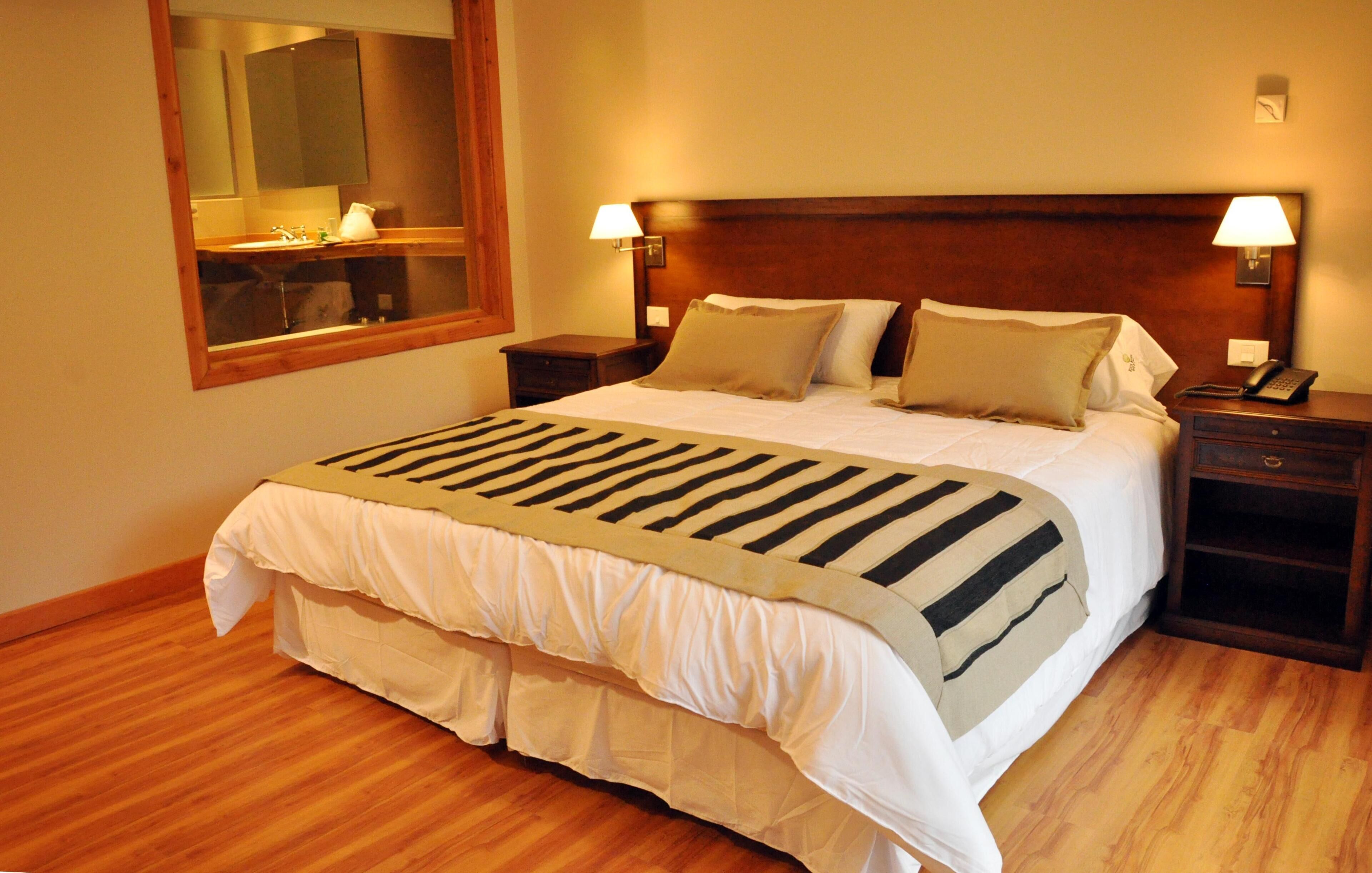 A hotel room with a large bed, wooden headboard, bedside lamps, a mirror, and a wooden floor.