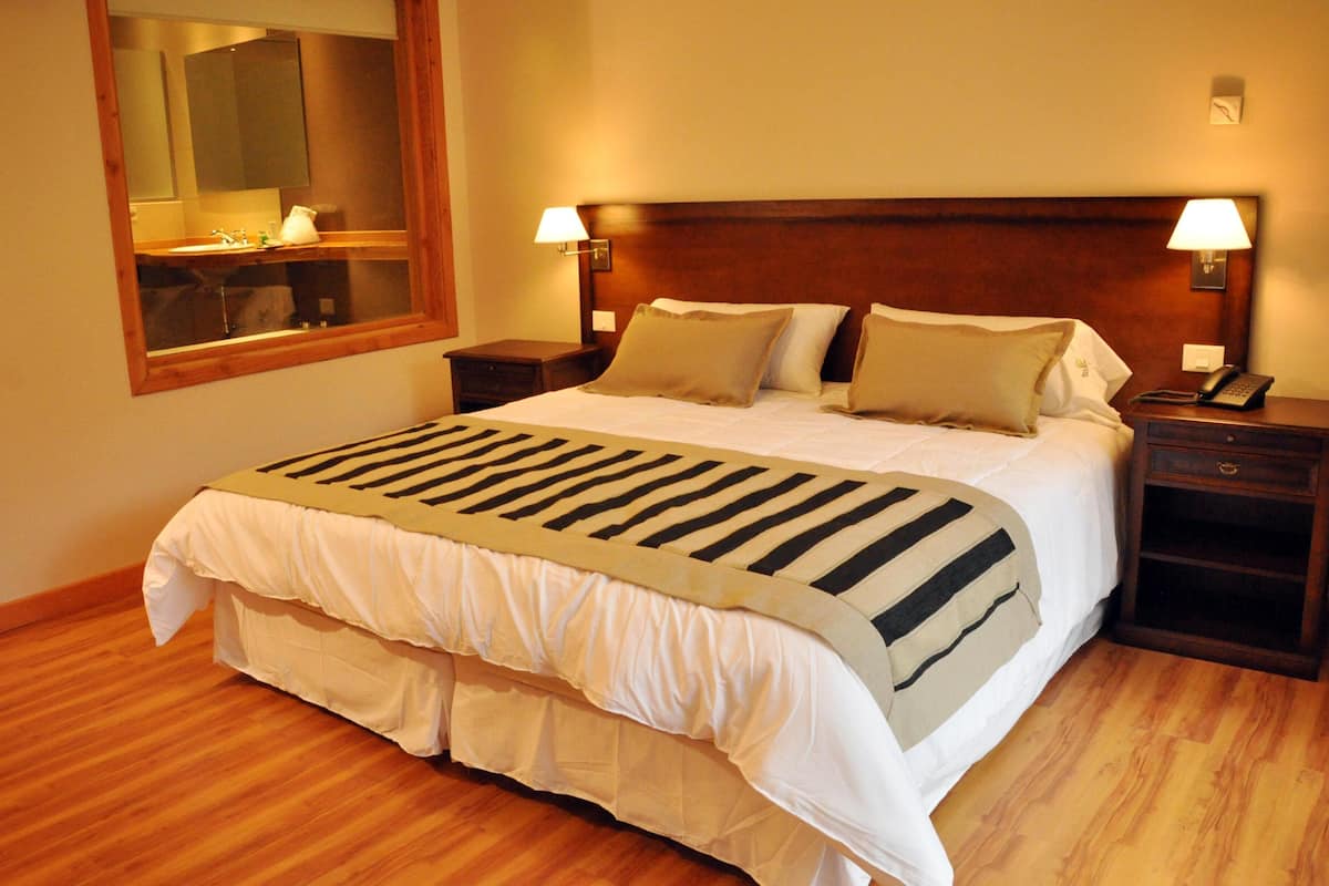 A hotel room with a large bed, wooden headboard, bedside lamps, a mirror, and a wooden floor.