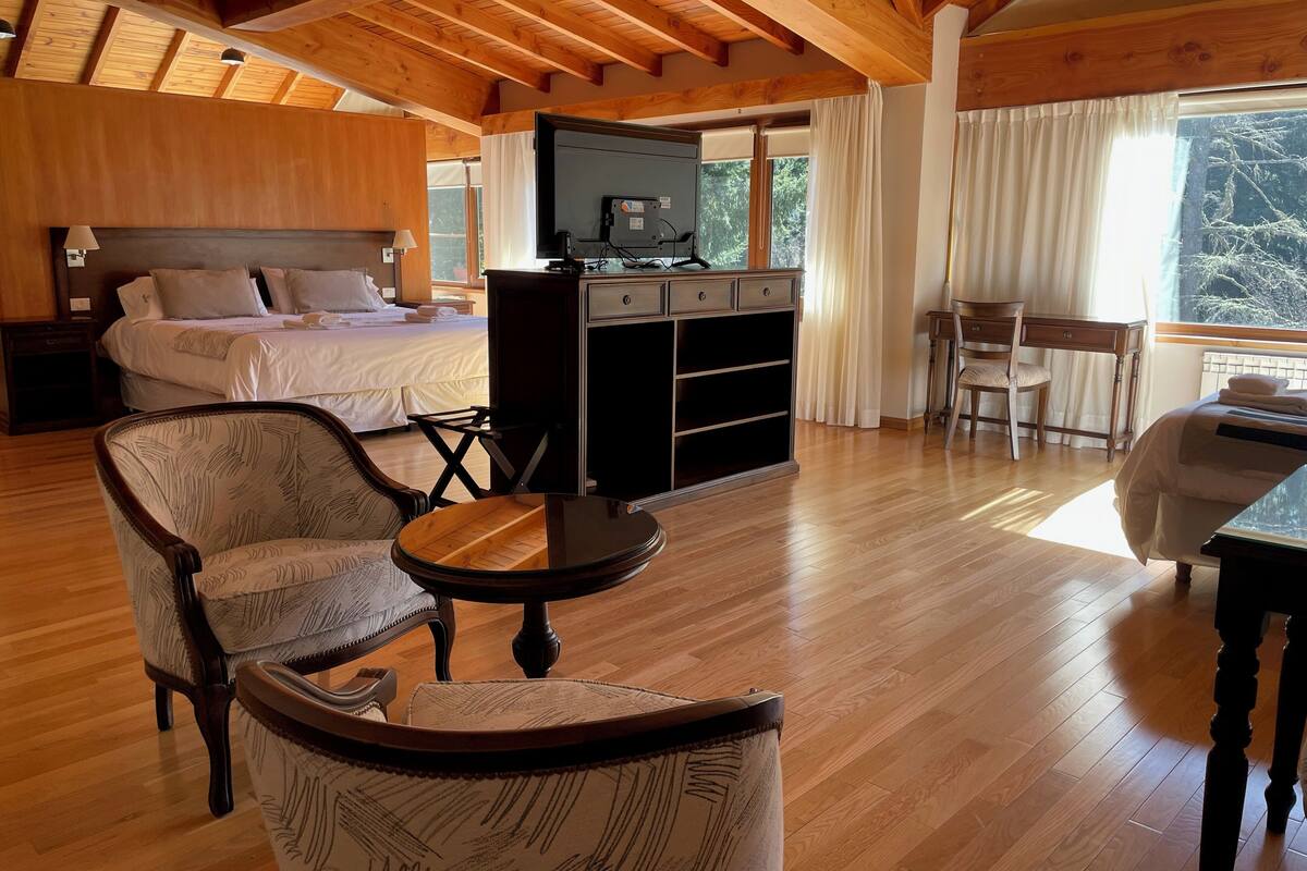 A spacious room with a wooden ceiling, a bed with white bedding, a television on a wooden cabinet, and a dining area with a chair and table.