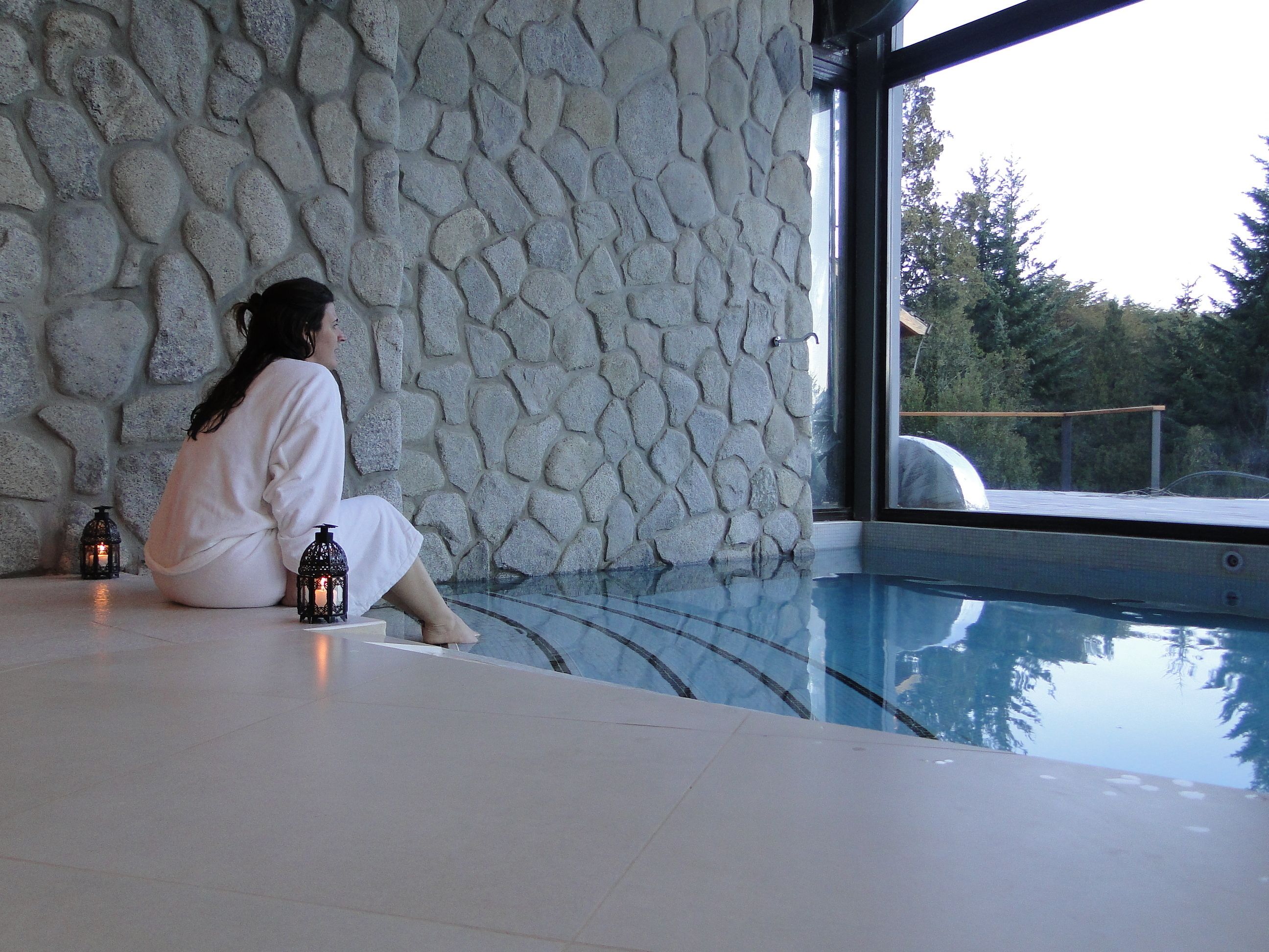 Una persona sentada junto a un muro de piedra, al lado de una piscina con vistas a árboles.