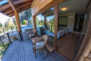 Una terraza de madera con mobiliario de mimbre y un ventanal que da a una habitación con suelo de madera y una cama.