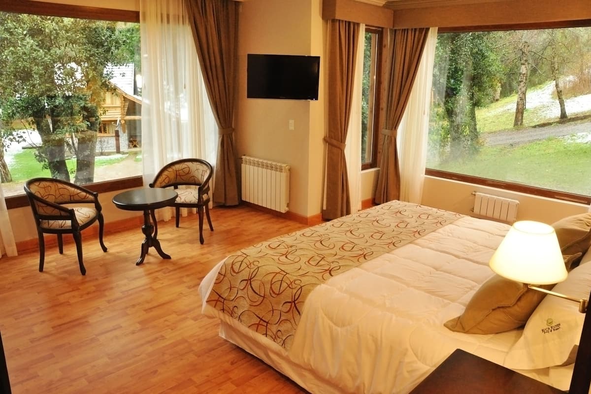A hotel room with a large bed, a TV mounted on the wall, a small table with two chairs, and large windows offering a view of the outdoors.