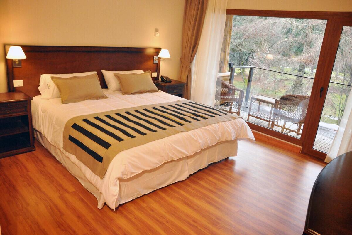 A large bed with a striped blanket, wooden headboard, two bedside lamps, and a view of outdoor furniture through a glass door.