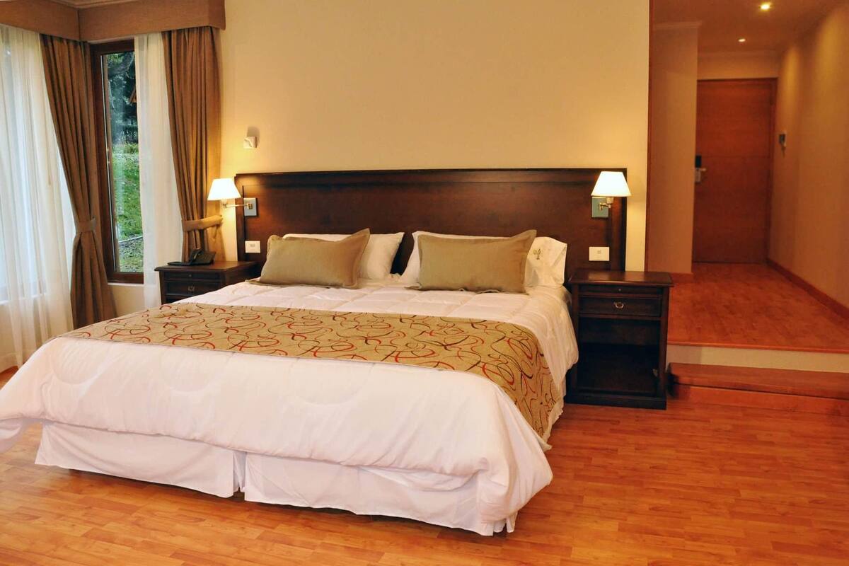 A hotel room with a large bed, two bedside tables with lamps, a wooden floor, and a view of a hallway.