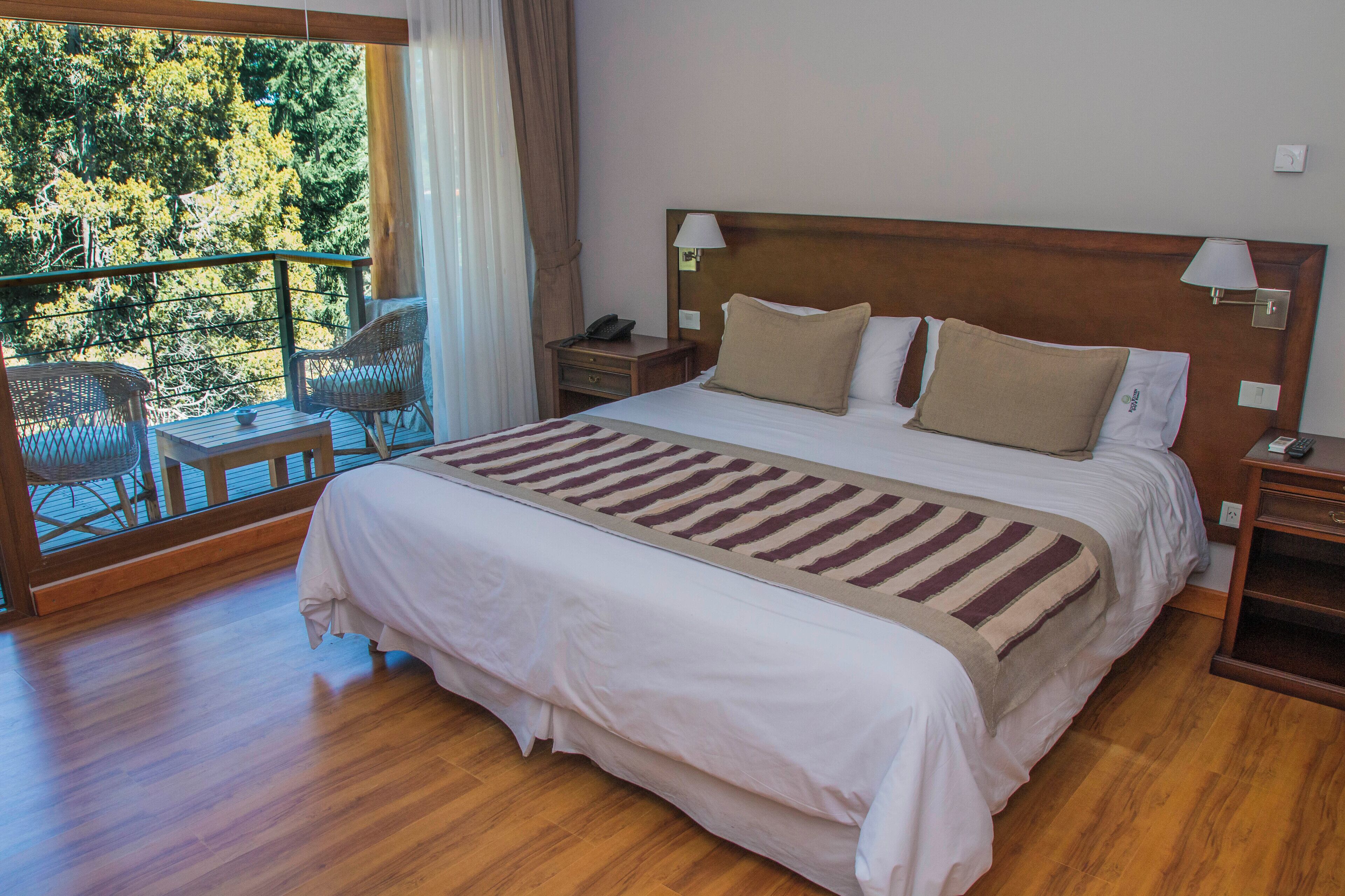 A spacious bedroom with a large bed, wooden floor, a balcony with wicker chairs, and a view of trees.