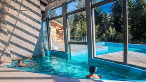 A swimming pool with clear blue water, surrounded by large windows offering views of a forested area. - Ruca Kuyen Golf & Resort (Villa La Angostura)