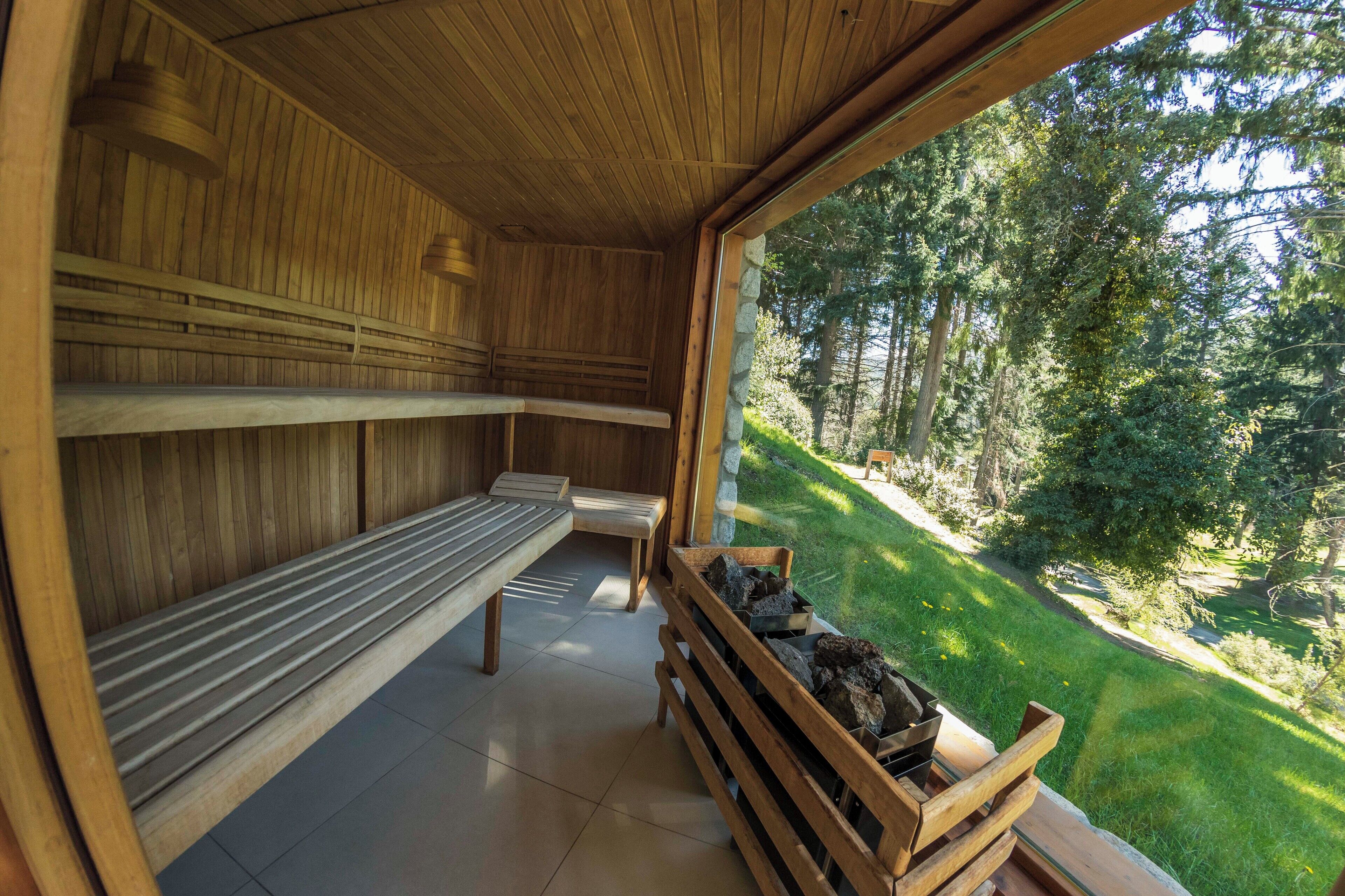 A sauna room with wooden benches and a stone bench, a stone wall, and a view of a forested area.