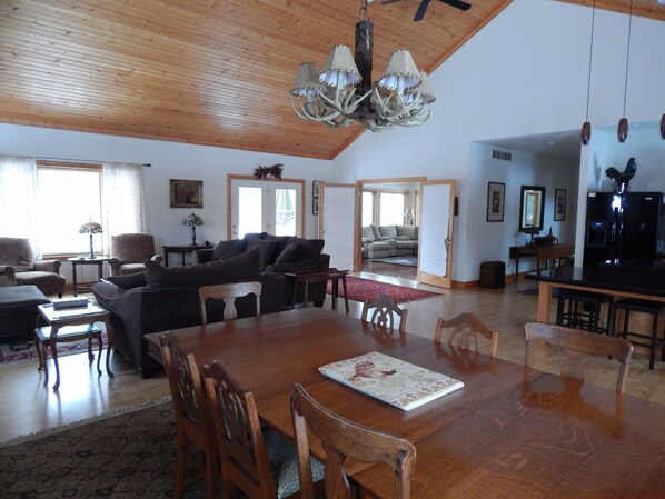 A spacious living area with a wooden ceiling, a chandelier, a dining table with chairs, a sofa, and a kitchen area with a refrigerator.