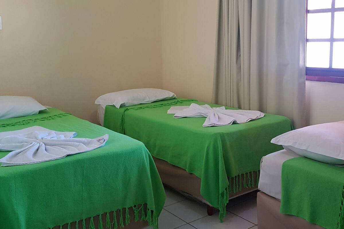 Una habitación con tres camas, cada una con cubrecama verde y almohadas blancas, y un ventanal con diseño de cuadrícula.