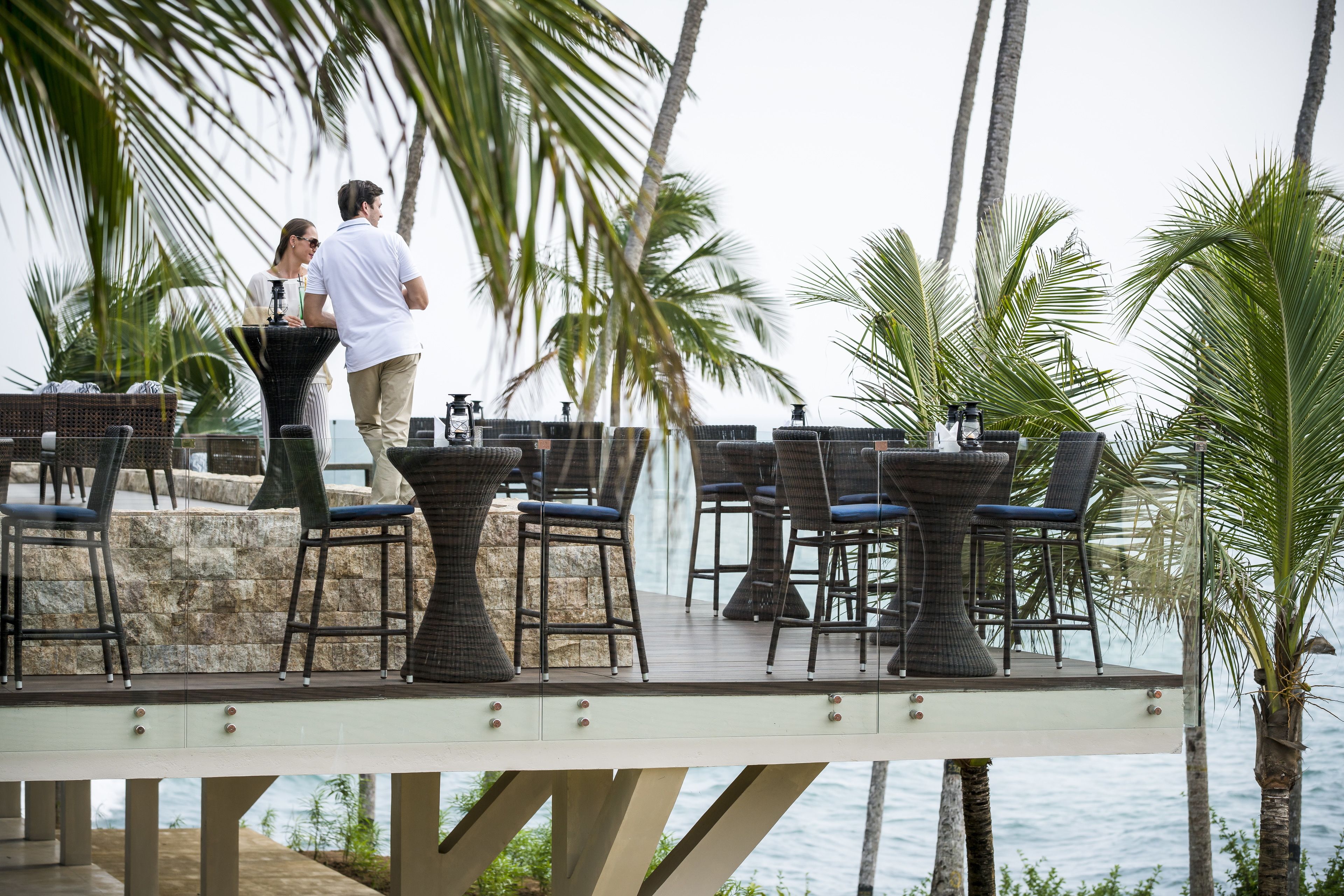 Un espace de restauration en plein air, avec des chaises et des tables en osier, donnant sur une étendue d’eau et entouré de palmiers.