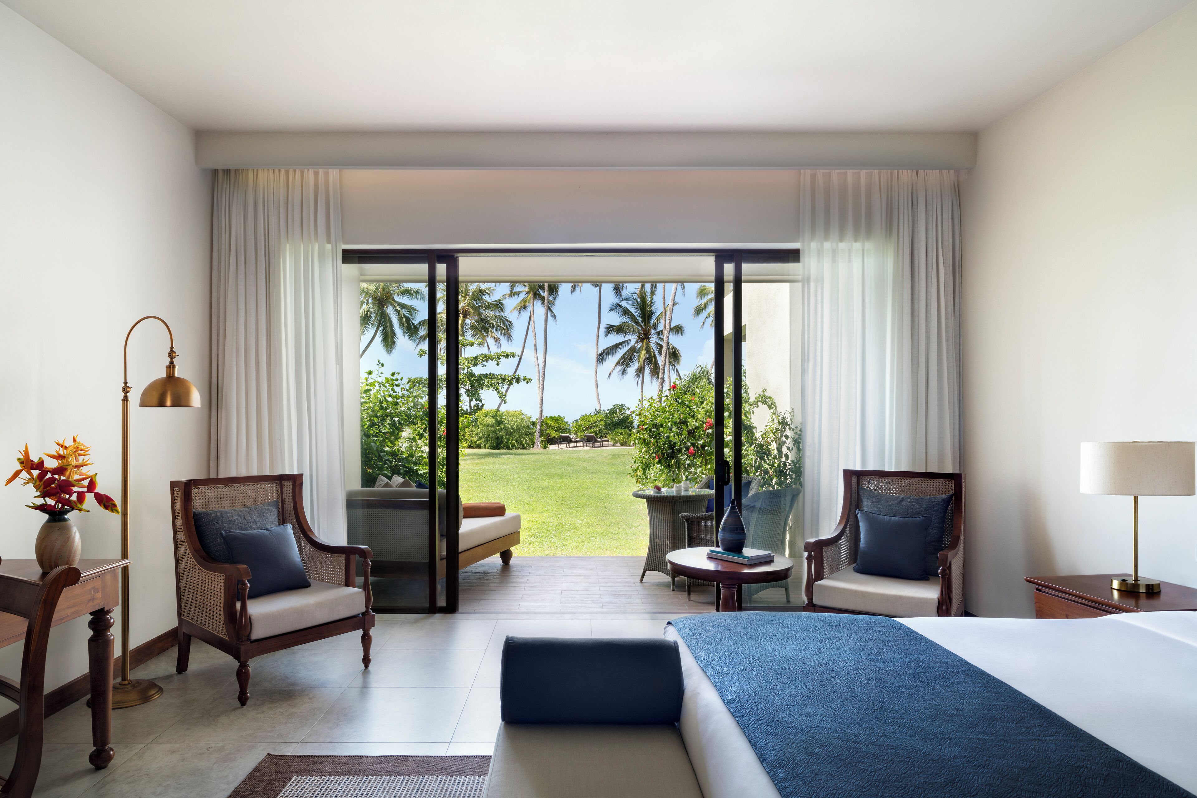 A hotel room with a large bed, two armchairs, a small table, and a view of a tropical garden through a sliding glass door.