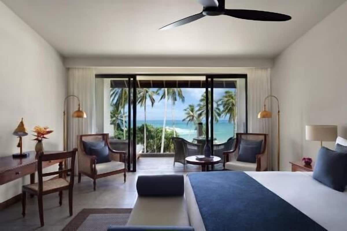 Habitación de hotel con cama, dos sillas, una mesa, ventilador de techo y vista al océano.
