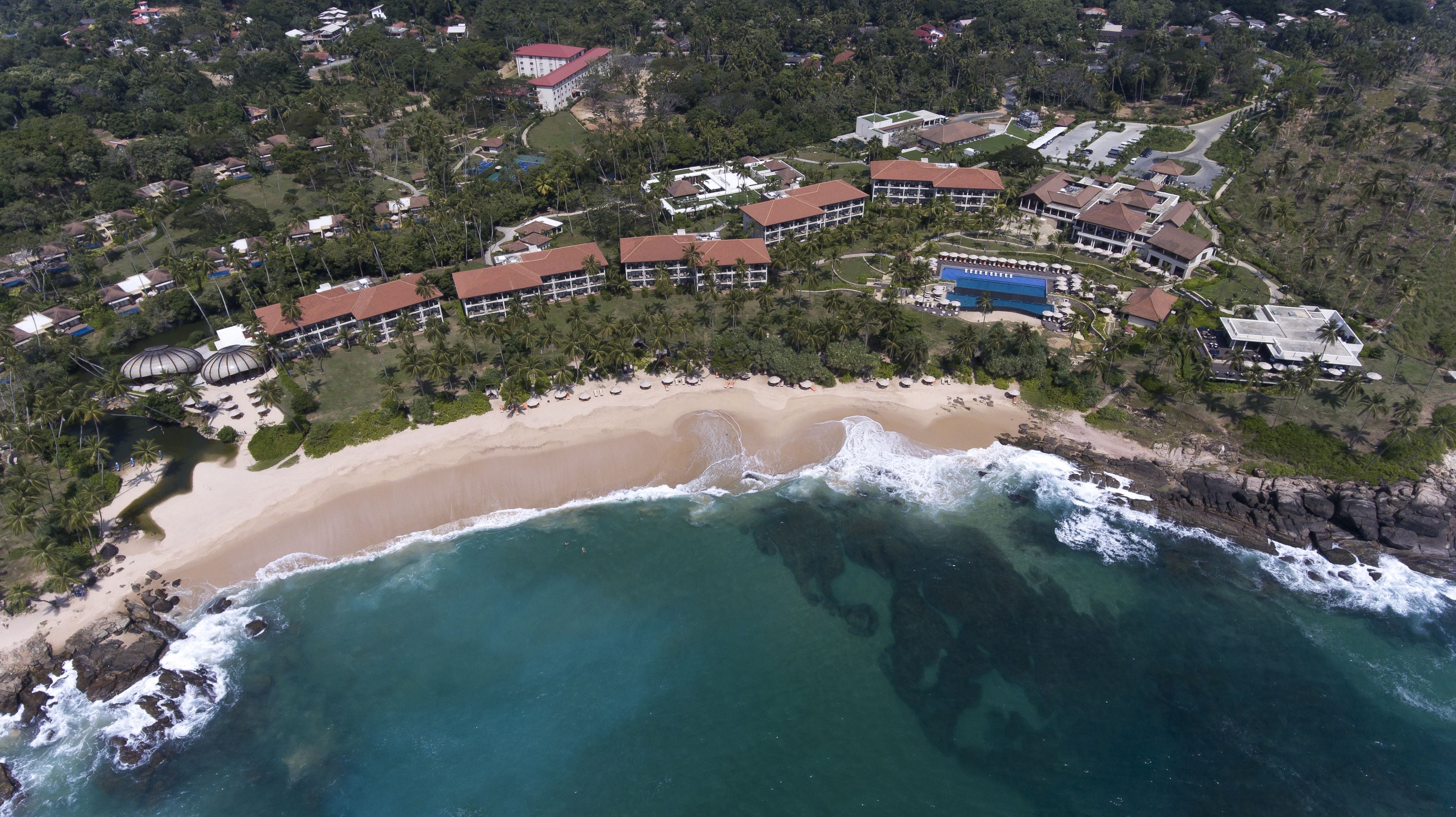 An aerial view of a coastal resort with beachfront villas, a sandy beach, and a rocky shoreline.