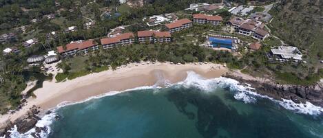An aerial view of a coastal resort with beachfront villas, a sandy beach, and a rocky shoreline.