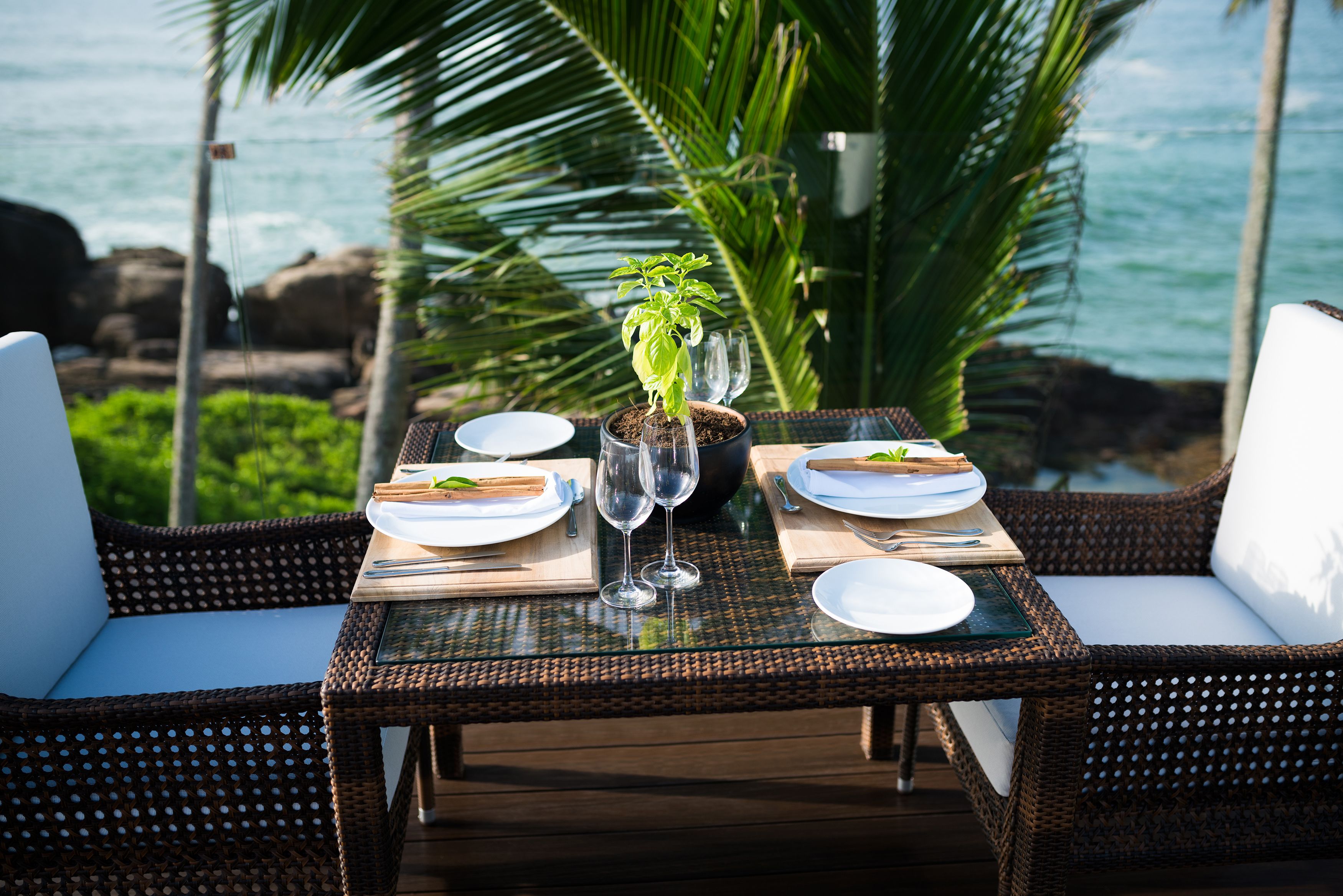A dining table set for two with white plates, cutlery, and wine glasses, surrounded by wicker chairs with white cushions, overlooking a rocky coastline.
