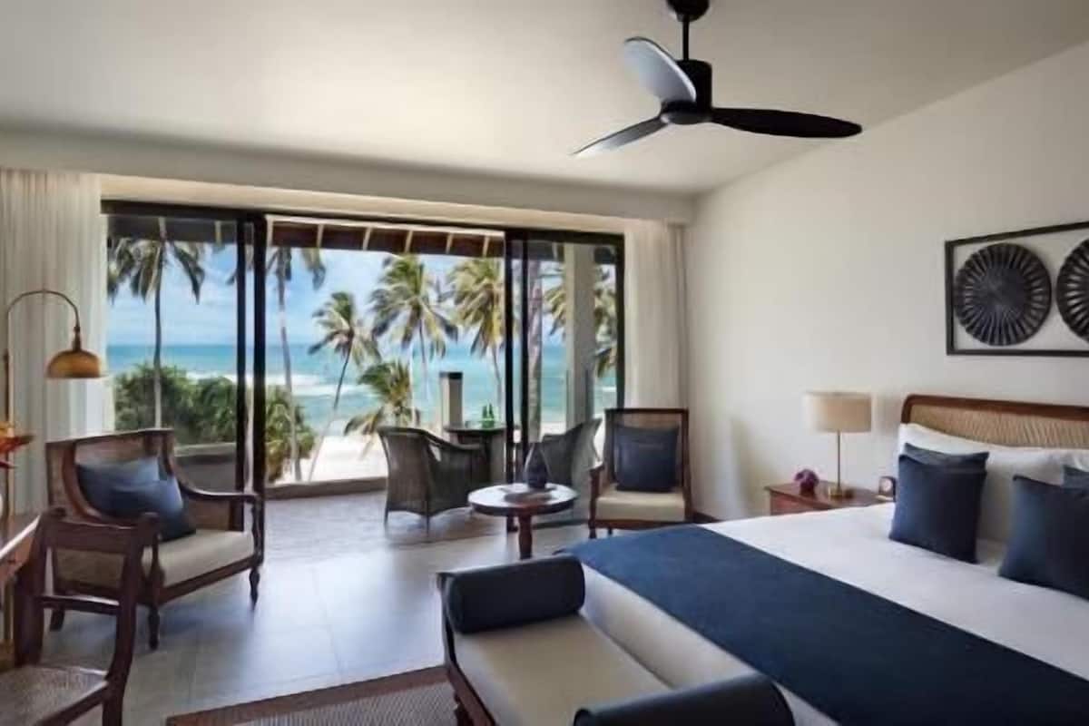Una habitación de hotel con cama, ventilador de techo, vista a la playa y un ventanal grande.