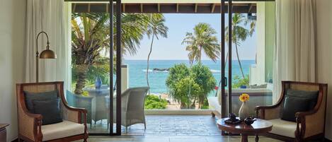 A room with a bed, two wicker chairs, a small table with a vase, and a view of the ocean and palm trees through large sliding glass doors.