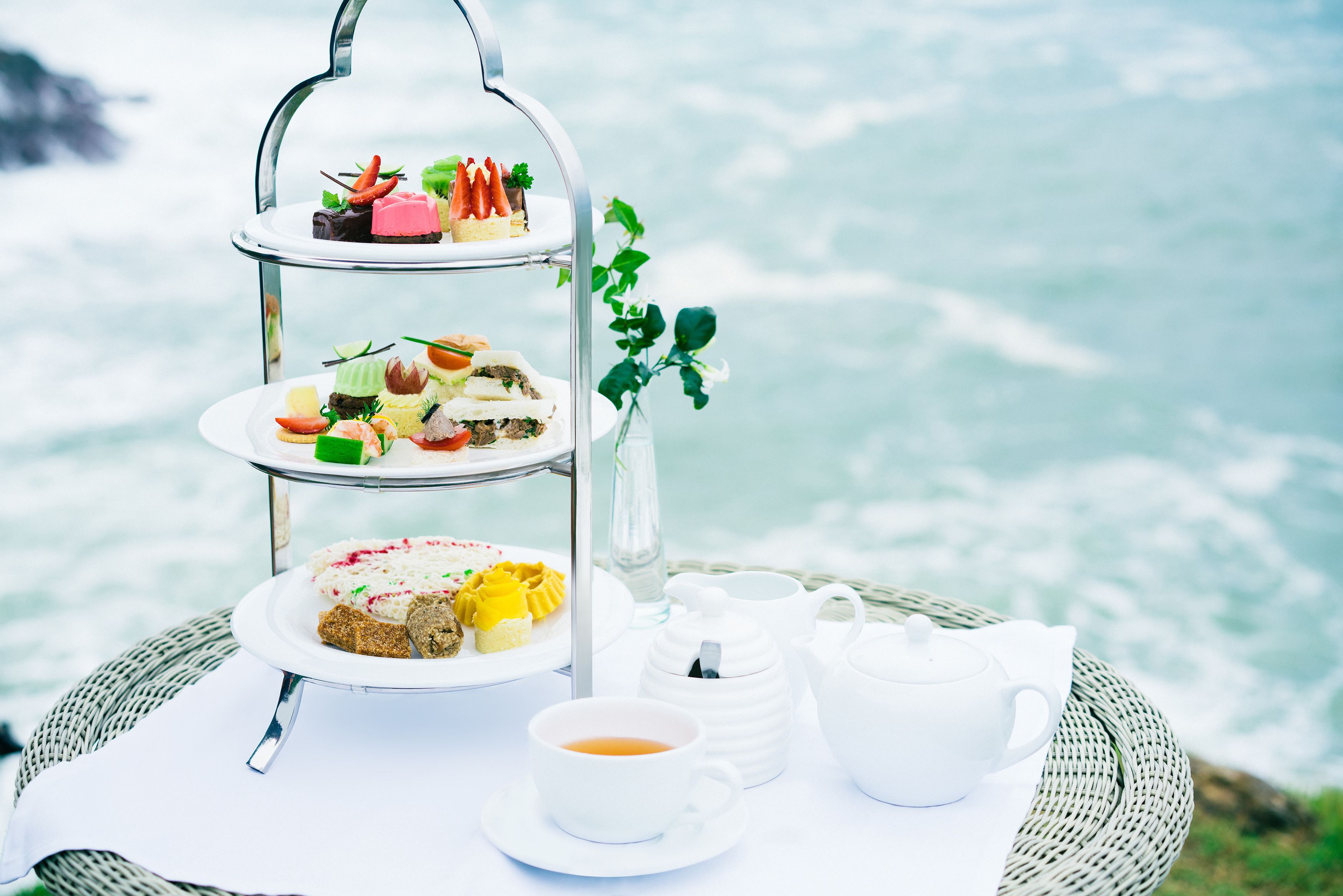A tiered tea service set on a wicker table with a view of the ocean.