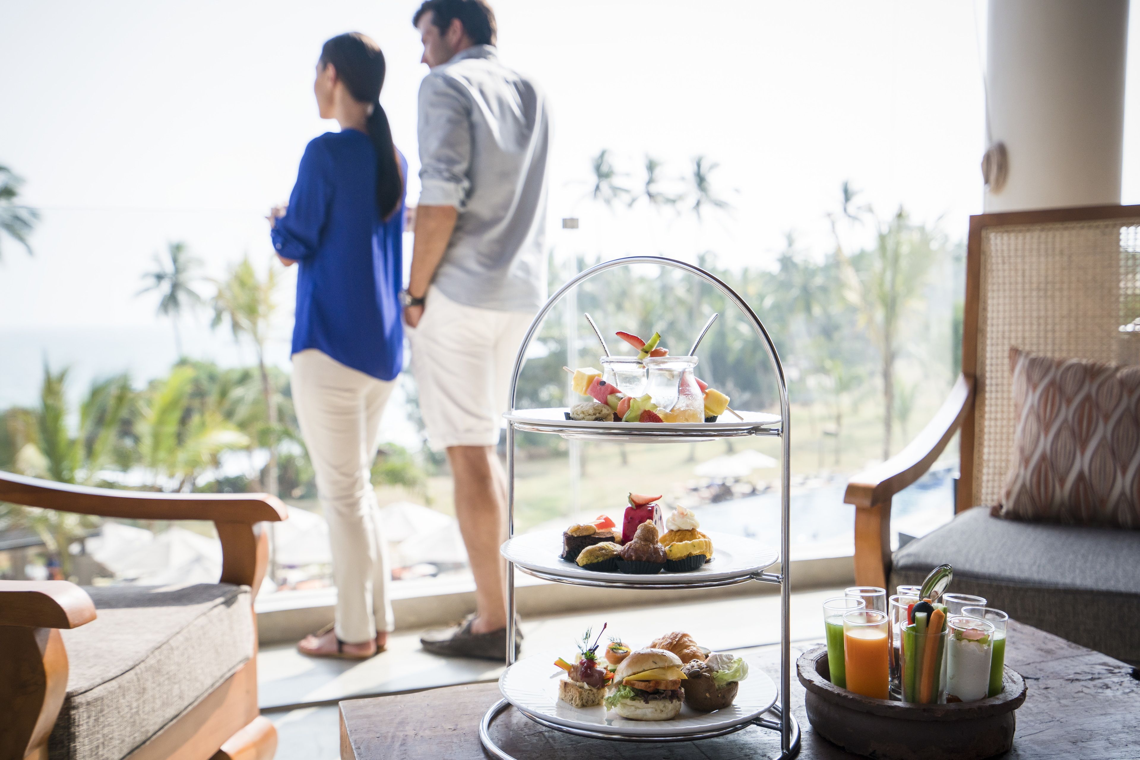 A tiered dessert stand with various pastries and a beverage tray on a balcony overlooking a tropical landscape.