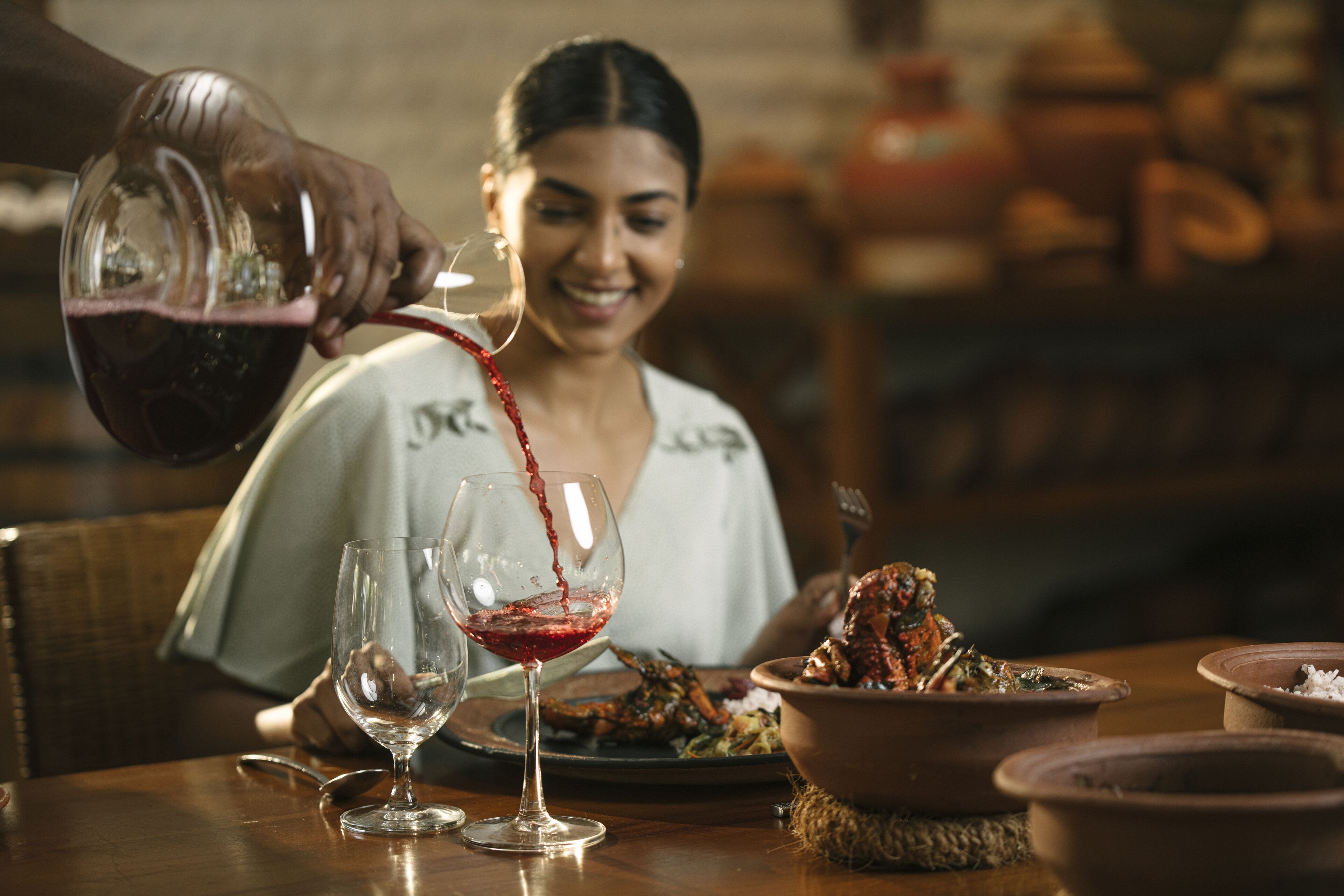 A dining table with a person pouring wine into a glass, a bowl of food, and a spoon.