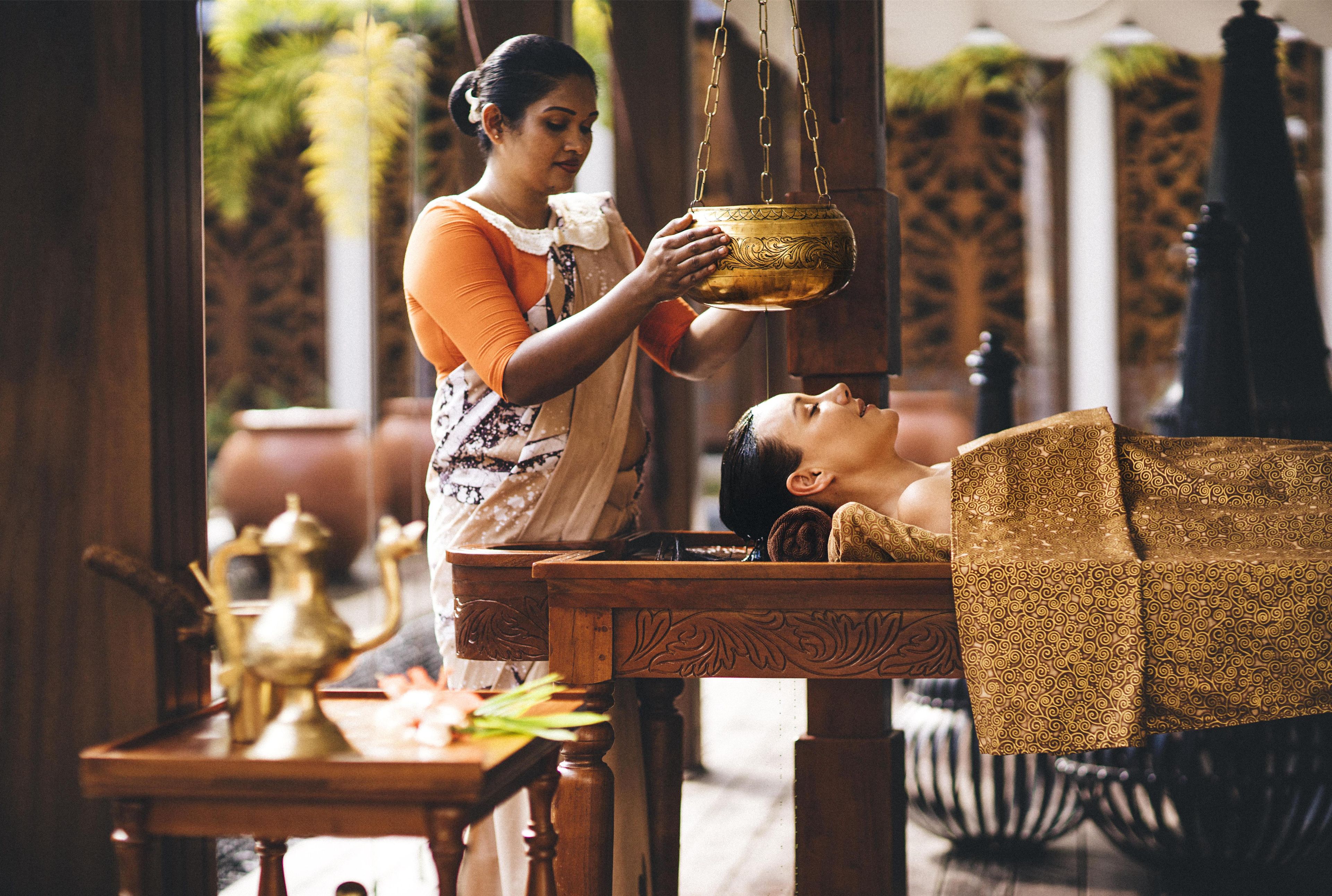 A person performing a ritual with a golden bowl, a table with a brass teapot, and a patterned cushion.