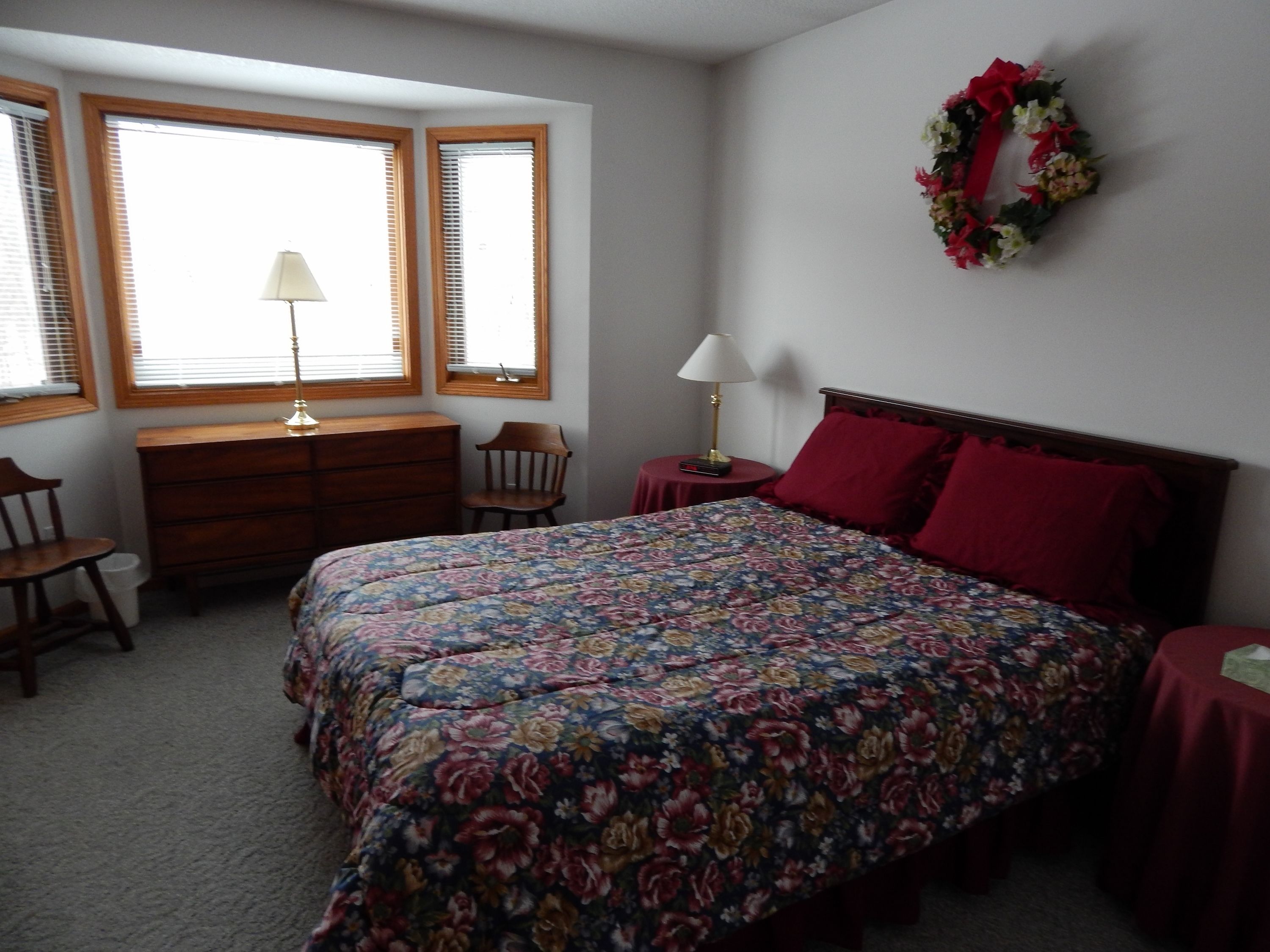 A bedroom with a floral bedspread, a wooden dresser, a chair, a nightstand with a lamp, and a wreath on the wall.