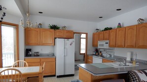 A kitchen with wooden cabinets, a refrigerator, a microwave, and a sink. There is a dining table with chairs and a trash can. - Augusta House (Galena)