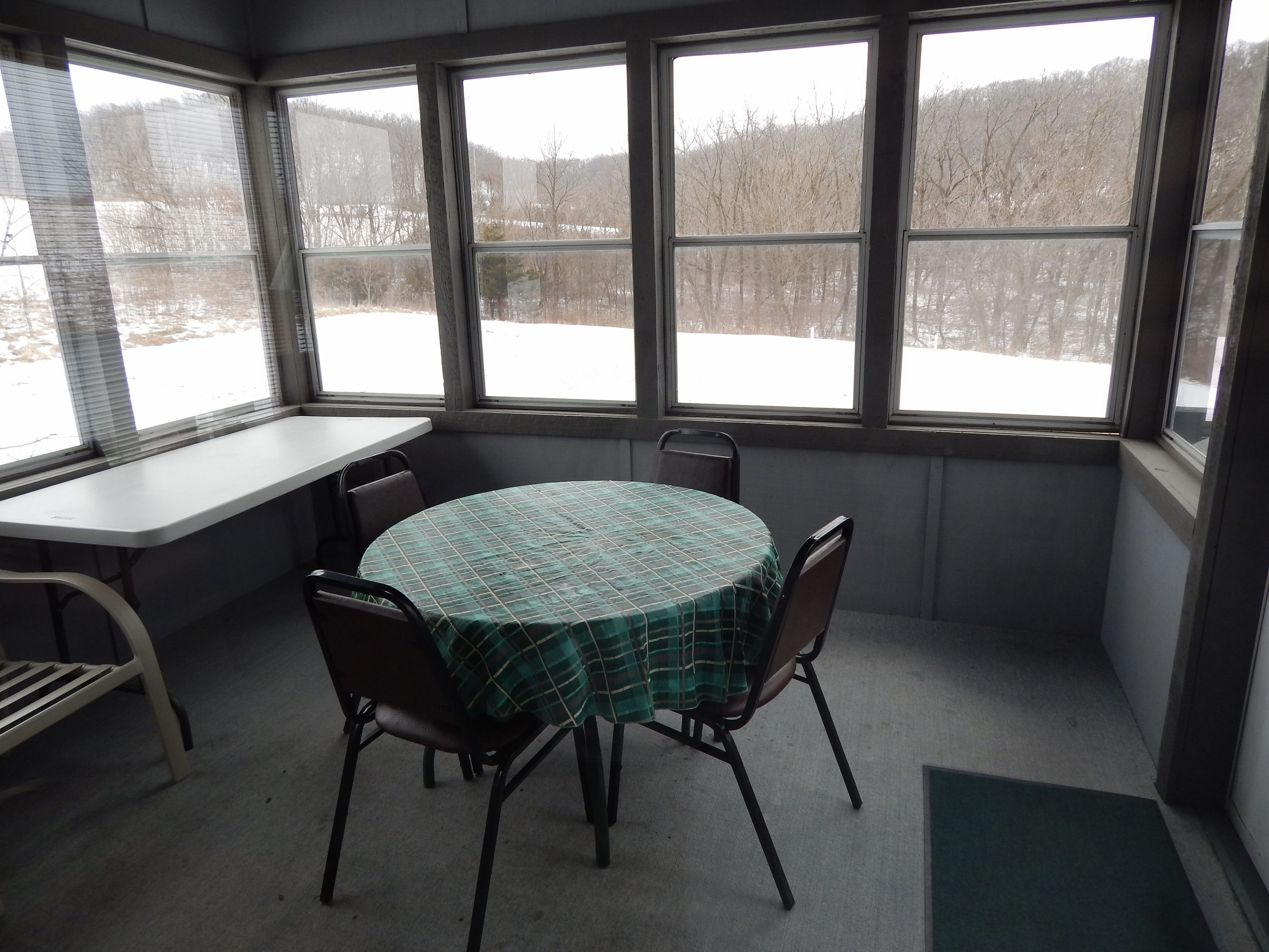 A room with a round table covered by a green checkered tablecloth, four chairs, a bench, and large windows offering a view of a snowy landscape.