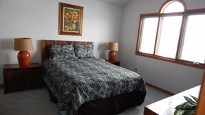 A bedroom with a bed, two nightstands, two lamps, a framed floral painting, a window with blinds, and a plant. - Augusta House (Galena)