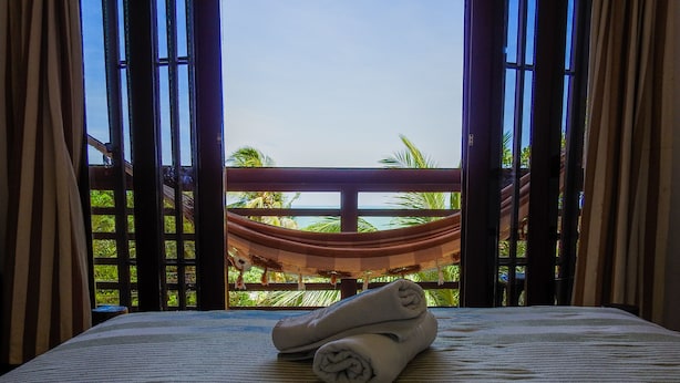 A bed with a striped bedspread and a rolled towel, a hammock in the background, and a view of palm trees through a window with curtains. - Apart Hotel Casa Grande (Natal)