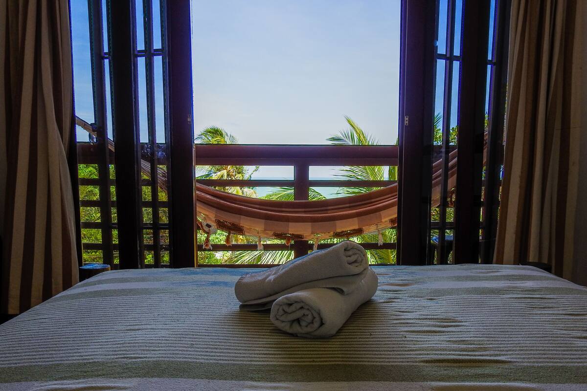 A bed with a striped bedspread and a rolled towel, a hammock in the background, and a view of palm trees through a window with curtains.