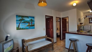 A room with a wooden bench, a painting of a tropical scene, a kitchen area with stools, and a door leading to another room. - Apart Hotel Casa Grande (Natal)