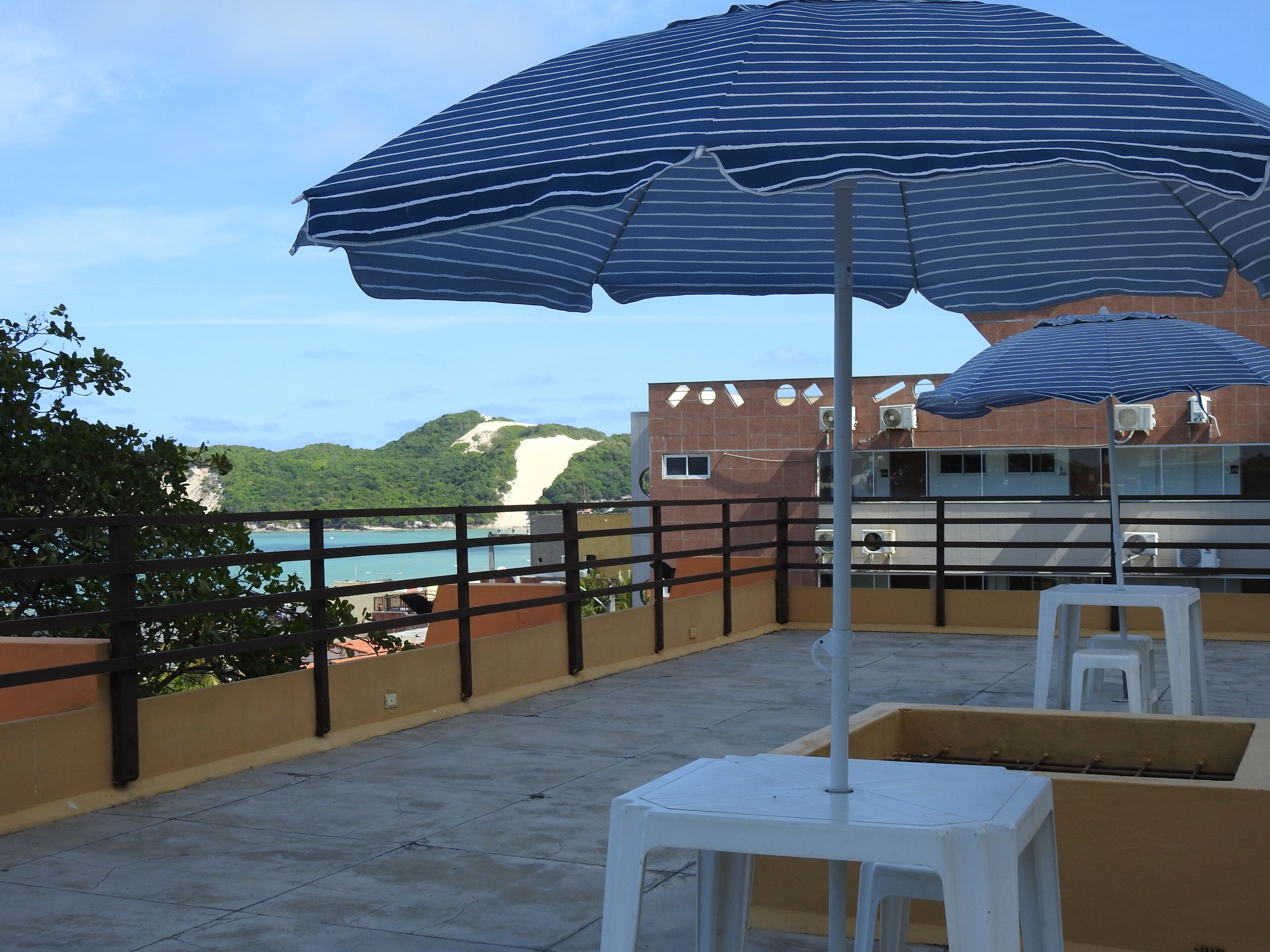 A terrace with a blue-striped umbrella, white chairs, and a view of a beach and mountains.