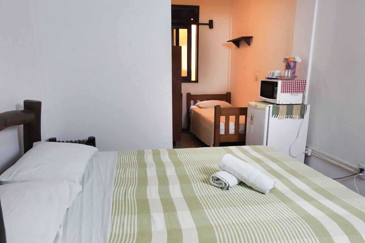 A small, cozy room with a single bed, a wooden headboard, a small desk with a computer, and a refrigerator.