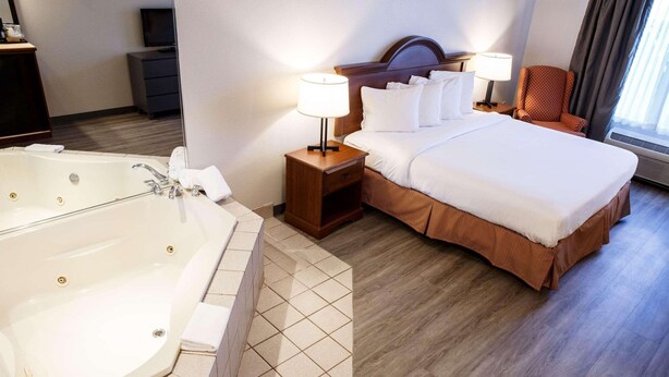 A hotel room with a large bathtub, a neatly made bed, a wooden nightstand with a lamp, and a chair by the window.
