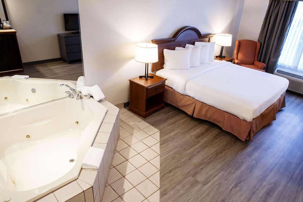 Une chambre d’hôtel avec une grande baignoire, un lit soigneusement fait, une table de chevet en bois avec une lampe et une chaise près de la fenêtre.