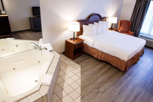 A hotel room with a large bathtub, a neatly made bed, a wooden nightstand with a lamp, and a chair by the window. - Country Inn & Suites by Radisson, Harrisburg West Mechanicsburg (Mechanicsburg)