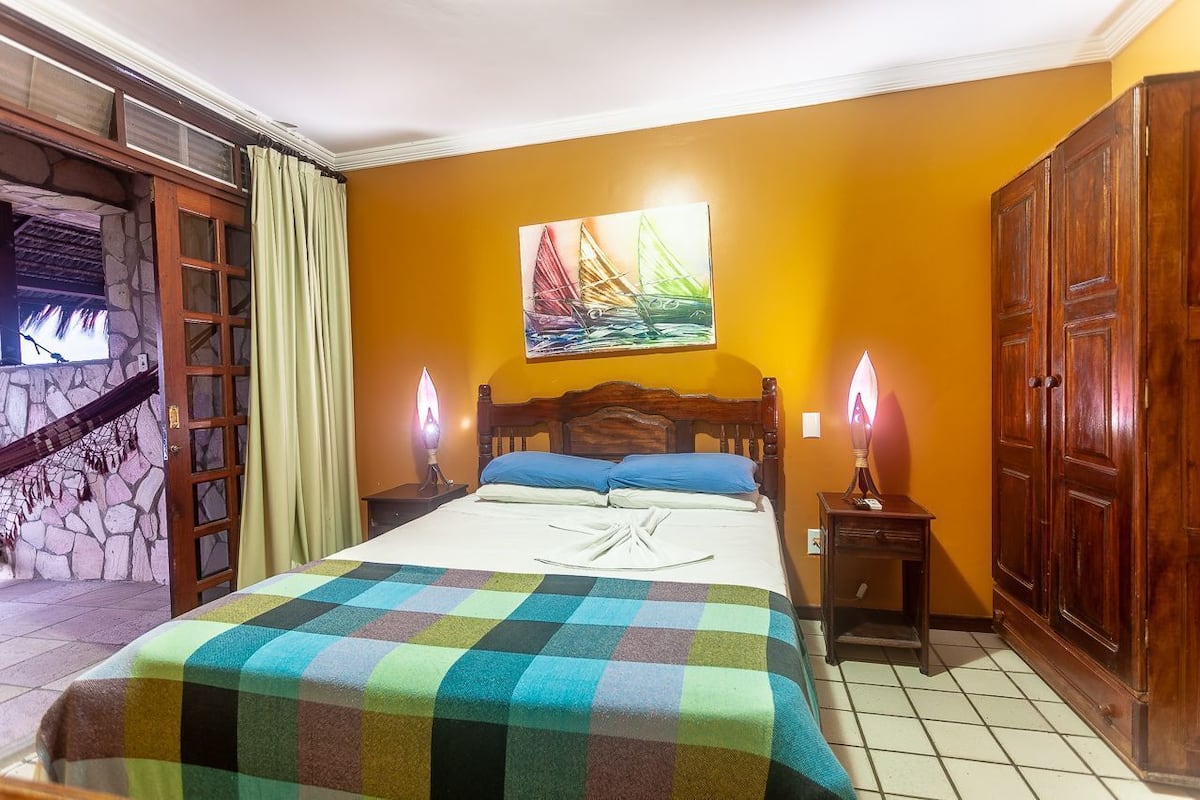 A hotel room with a bed, a painting, a wooden wardrobe, and a stone wall.