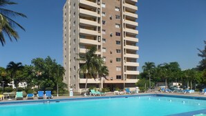 Une piscine entourée de chaises longues et de palmiers, le tout bordé d’un grand immeuble doté de nombreux balcons.