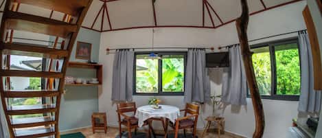 Un comedor con una escalera espiral de madera, una mesa redonda con sillas y vistas a vegetación a través de amplios ventanales.