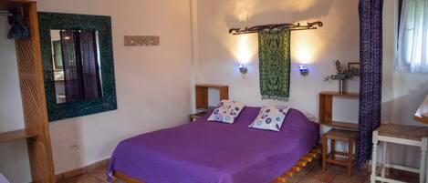 A bedroom with a bed, wooden headboard, a nightstand, a small table, a mirror, and a wall-mounted light fixture.