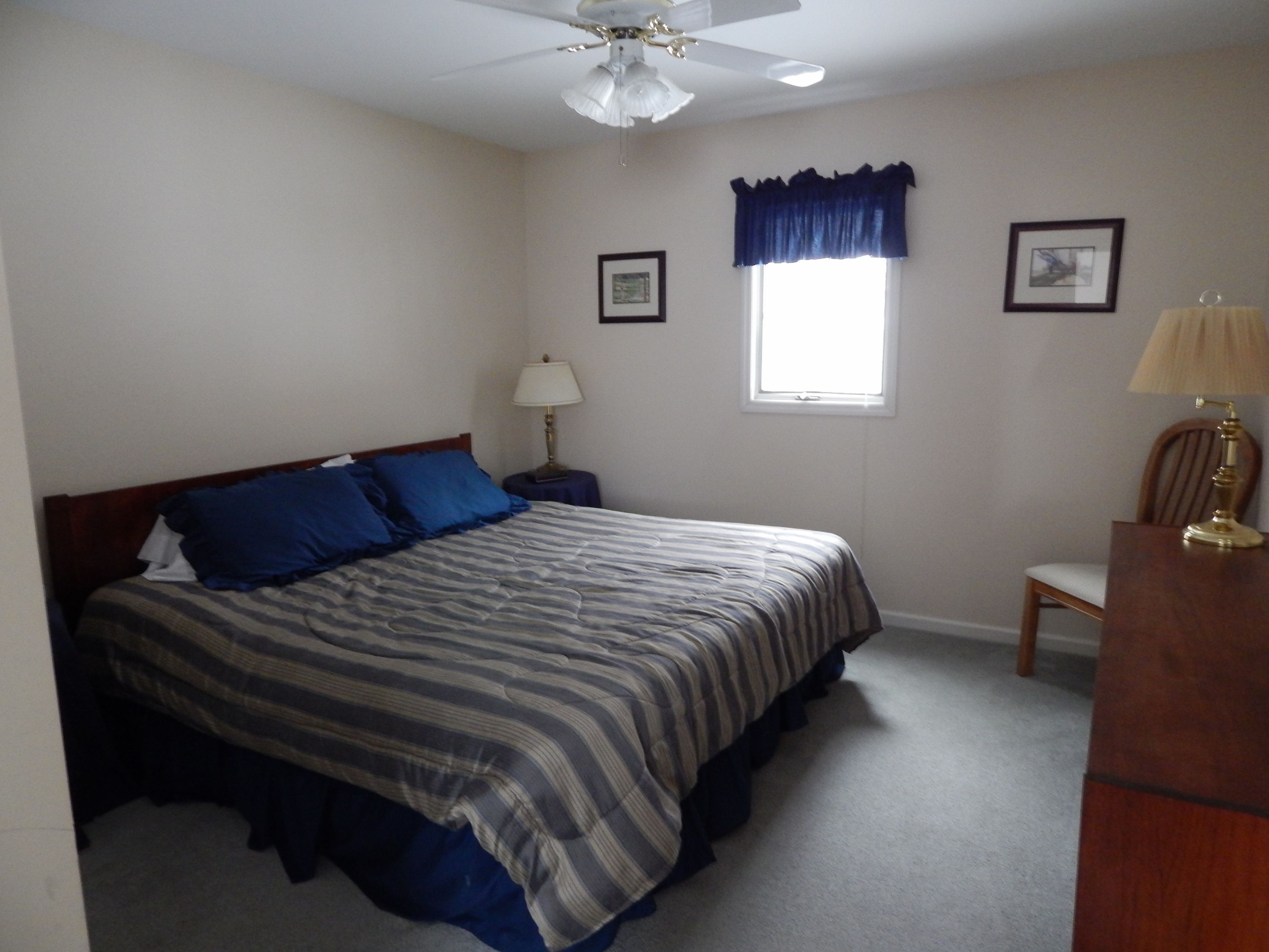 A bedroom with a bed, a nightstand, a desk, a chair, a window with curtains, a ceiling fan, and two framed pictures on the wall.