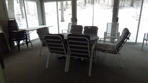 A covered outdoor dining area with a round table and striped chairs, large windows, and a view of a snowy landscape. - House on Pond Court (Galena)
