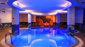 A large indoor swimming pool with a central island, surrounded by lounge chairs and a mural of a tropical scene. - Catamaran Quality Times - All Inclusive (Kemer)
