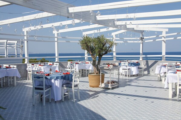 An outdoor dining area with white tables and chairs, a large tree, and a view of the sea.