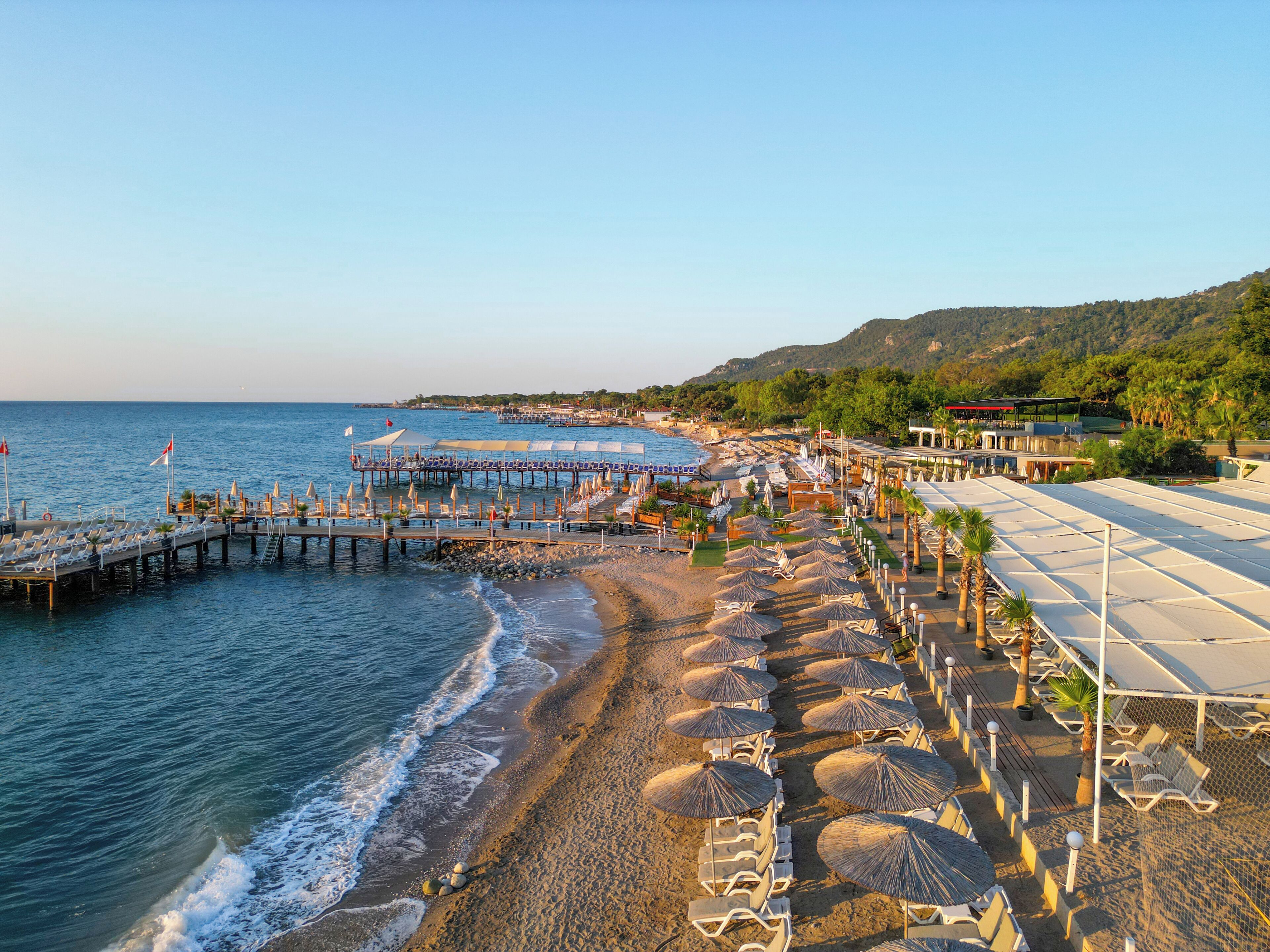 A beachside resort with rows of sun loungers and umbrellas, a pier extending into the sea, and a mountainous backdrop.