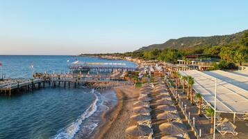 A beachside resort with rows of sun loungers and umbrellas, a pier extending into the sea, and a mountainous backdrop.