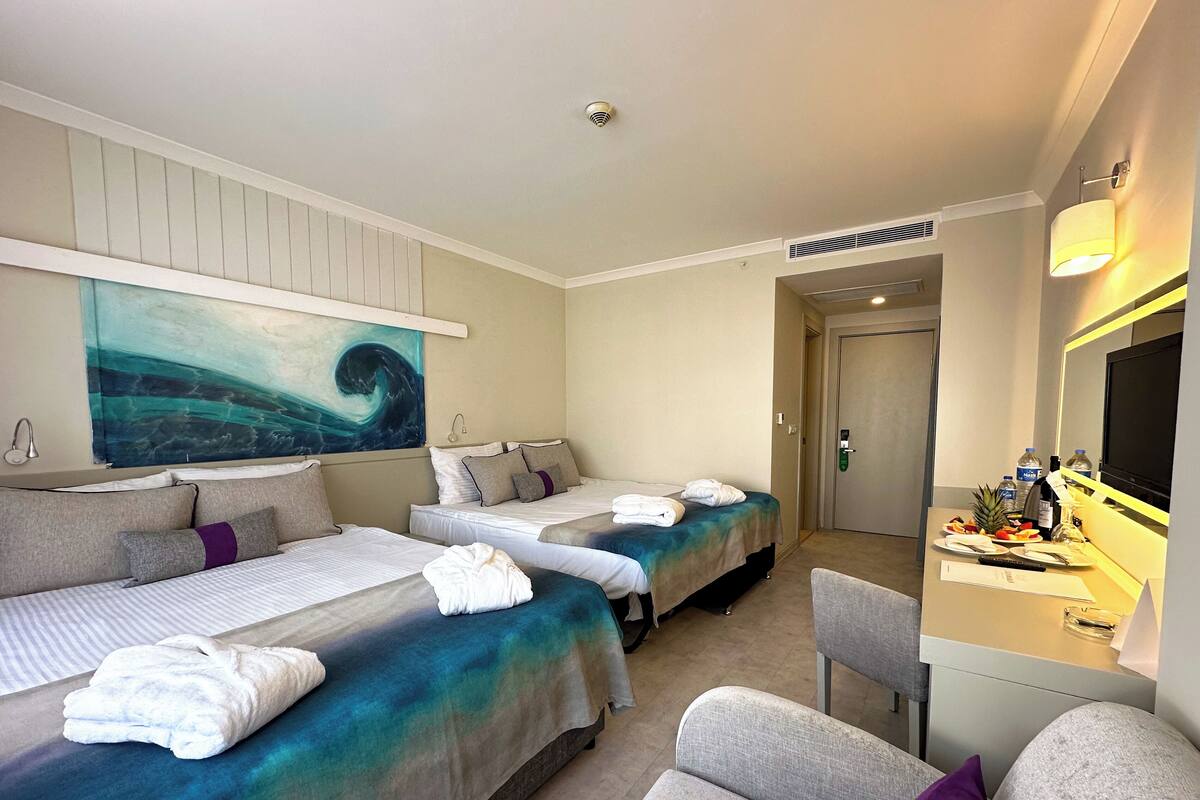 A hotel room with two beds, a desk with a mirror, and a painting of a wave on the wall.